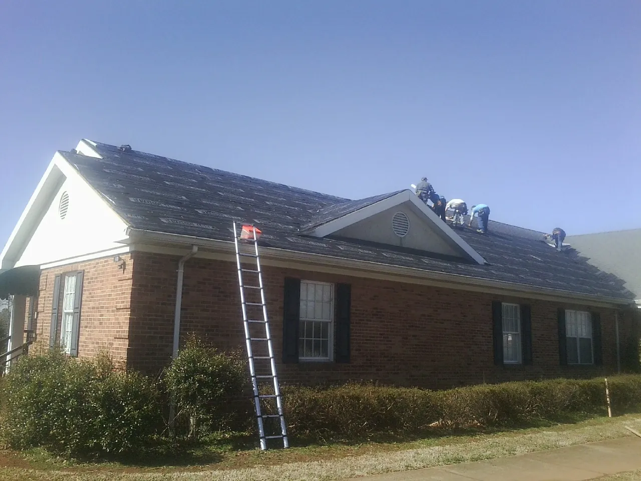Crew tearing shingles off a brick ranch roof with a ladder set — Metal Roof Repair work by our crew in Blissfield